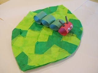 A caterpillar craft made from rings of construction paper connected together sitting on top of a leaf made from green tissue paper squares