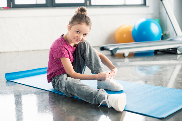 A little girl is seen tying her shoe while sitting on a mat.