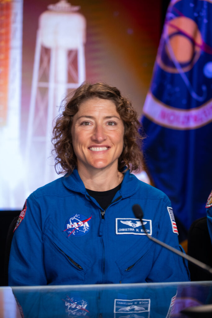 A young woman wearing an astronaut suit is shown smiling at the camera from the waist up.