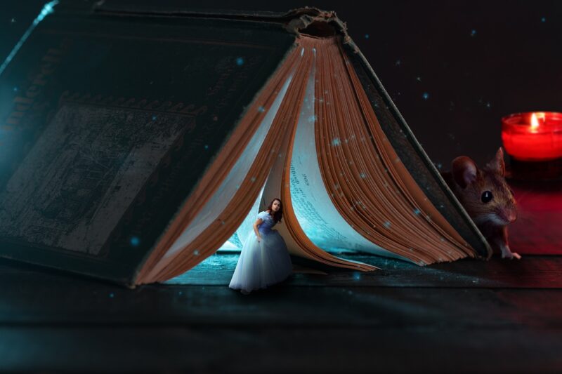 Woman dressed in a blue ball gown peering out of a book lit from inside, with a mouse nearby