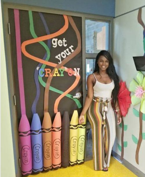 Teacher standing by a classroom door decorated with 3D paper crayons. Text reads "Get your cray-on."
