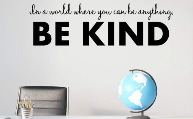 Vinyl letters on a classroom wall behind a teacher's desk saying In a world where you can be anything, be kind.