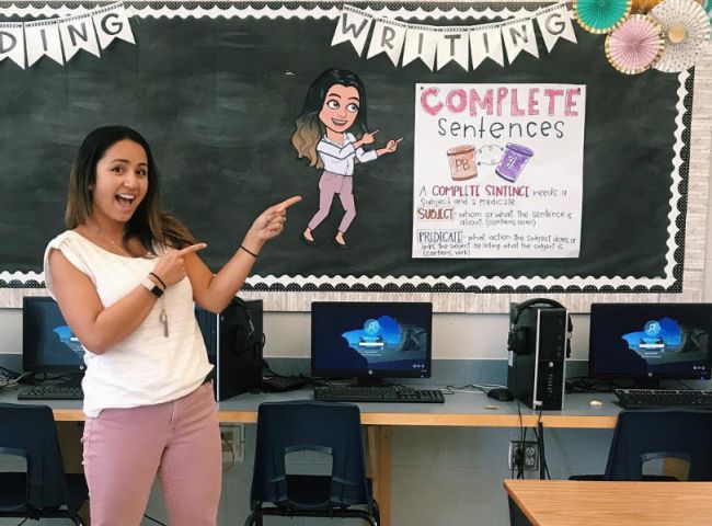 Teacher posing by a large Bitmoji-style poster of themselves pointing to the classroom rules