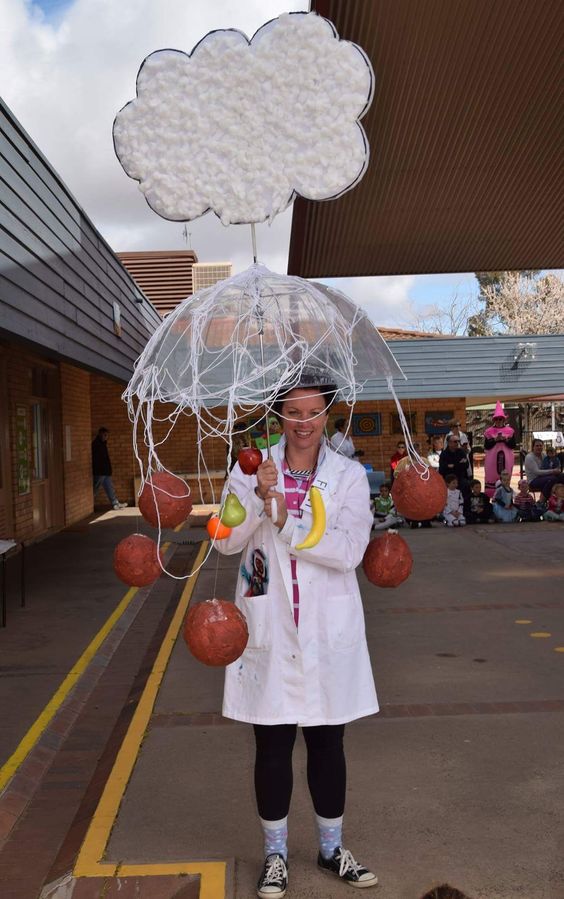 A woman is wearing a white lab coat and holding a clear umbrella that has a cloud coming out of it. There are meatballs and other food items hanging from the umbrella.