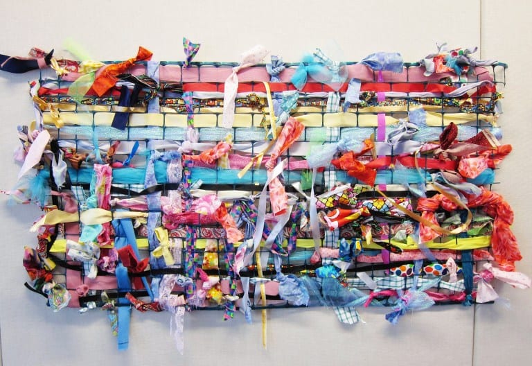 A colorful wall hanging made from plastic fencing and colorful strips of cloth