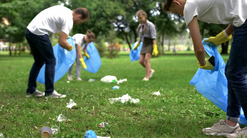 kids and adults wearing yellow plastic gloves gathering trash in a grassy park into blue trash bags