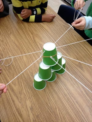 Students gathered around a table, forming a pyramid of green paper cups using only strings 