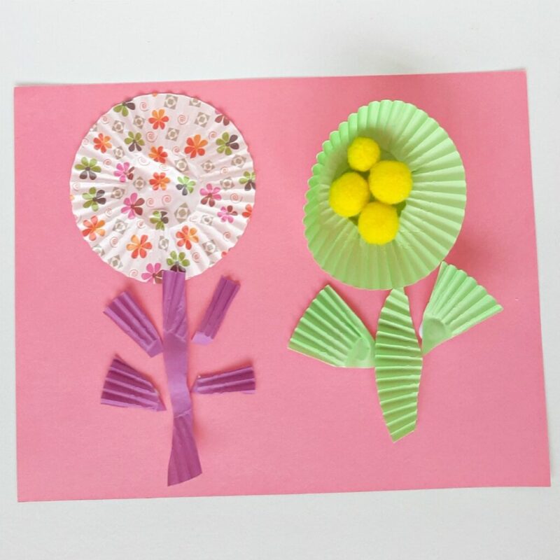 Two flowers are shown made from cut up cupcake liners and pom poms. They are on a pink piece of construction paper. Example of easy crafts for kids.