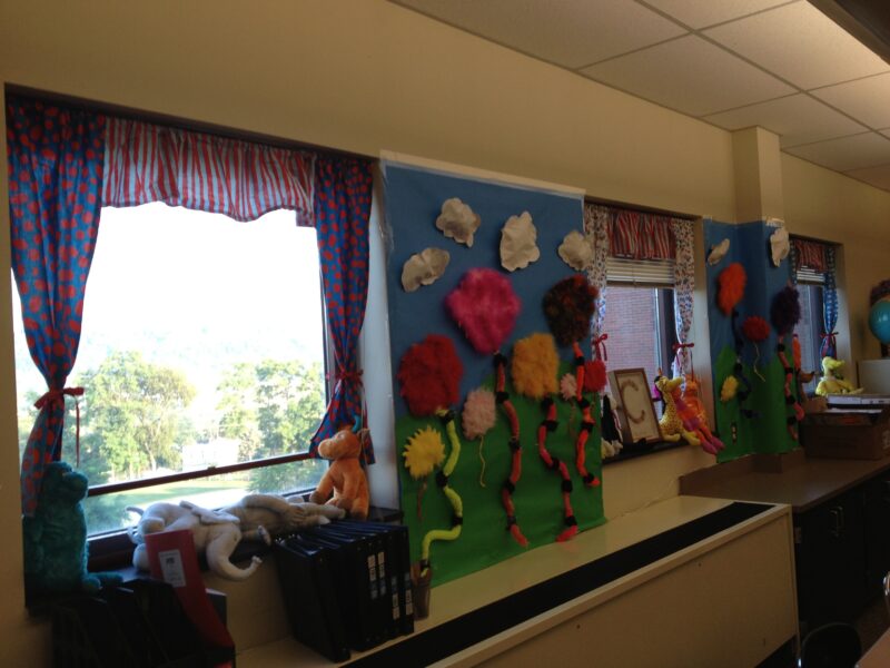 A bulletin board is bookended by some fun curtains on the windows of a classroom.