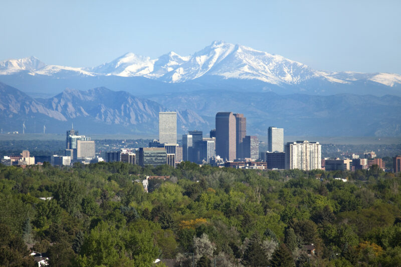 Snow covered Longs Peak, part of the Rocky Mountains stands tall in the background with green trees and the Downtown Denver skyscrapers as well as hotels, office buildings and apartment buildings filling the skyline, as an example of best family vacations