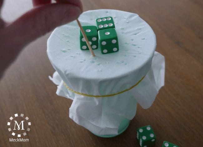 Green dice balanced on top of a cup covered with a tissue, while a student pokes holes with a toothpick (Dice Games)