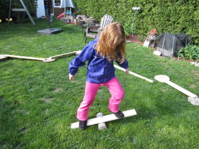 Child balancing on a plank of wood over a round piece, as part of diy obstacle courses for kids