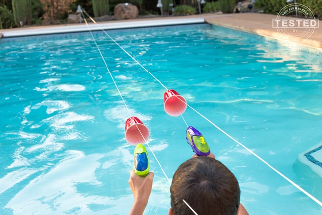 Kids using squirt guns to propel plastic cups along strings strung over a swimming pool, as an example of DIY obstacle courses for kids