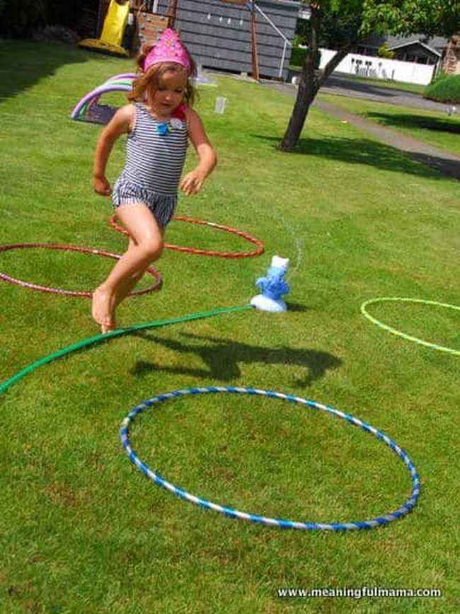 Little girl in a bathing suit running through a backyard obstacle course with sprinklers and other water features, as an example of DIY obstacle courses for kids