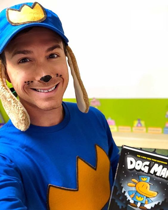 Book character costume ideas like this one shows a teacher wearing a blue hat with a yellow crown on it and dog ears as well as a blue sweatshirt with a yellow crown on it. The teacher's nose is painted black and he's holding the book Dog Man.