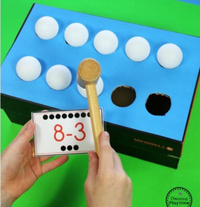 Student using a wooden mallet to tap ping pong balls into a cardboard box while holding a card saying 8-3