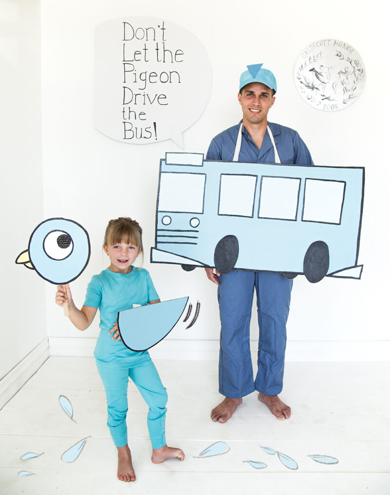A sign reads Don't let the pigeon drive the bus. a man is wearing blue and standing behind a cutout of a bus. A child is also dressed in blue and holding a pigeon head cut out.