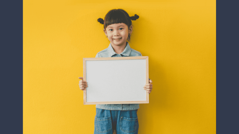 Young smiling girl standing in front of bright yellow wall holding a blank dry erase board