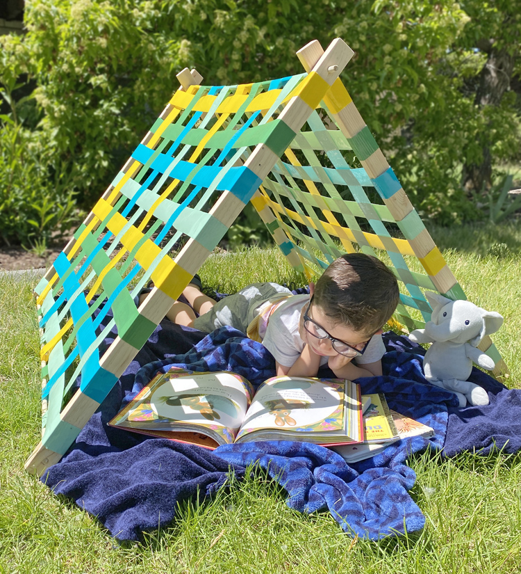 A boy reading reclines under a tent made from a wood frame and duct tape walls as an example of summer crafts for kids