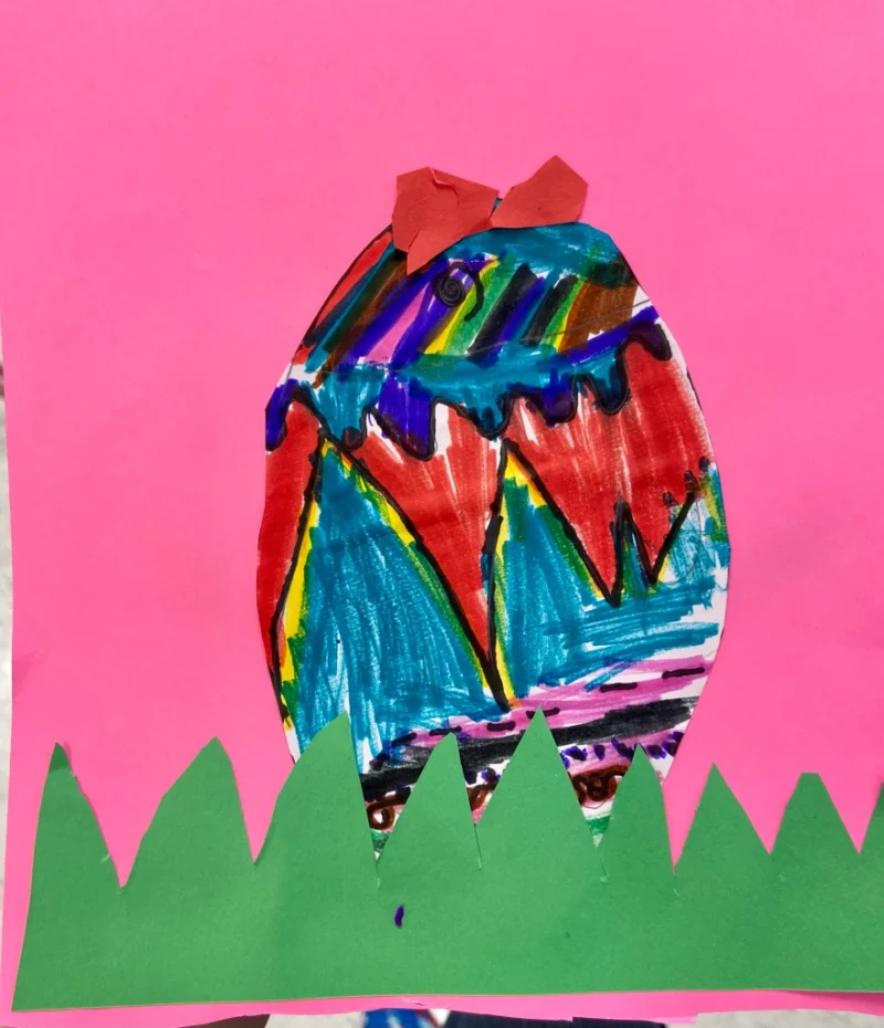 A colorful egg is drawn on a pink piece of construction paper with construction paper grass.