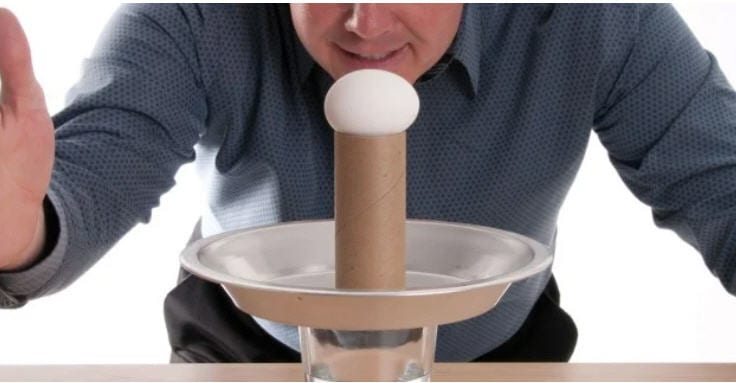 Egg on top of a toilet paper tube, standing on a plate on a glass of water, with a man ready to hit the plate