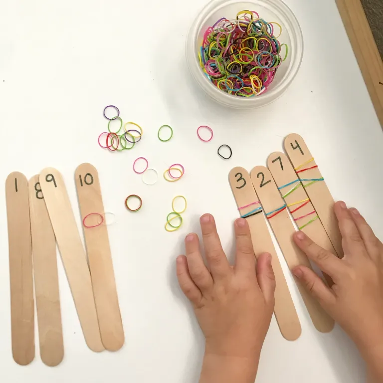 Popsicle sticks have numbers on the top of them and little hands are shown putting the correct number of elastics onto them (fine motor activities)