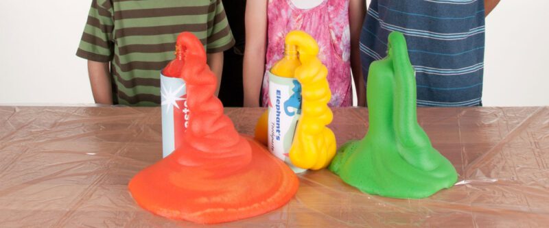 children are seen from the shoulders down standing behind three bottles that are overflowing with red, yellow, and green fluid. (easy science experiments)