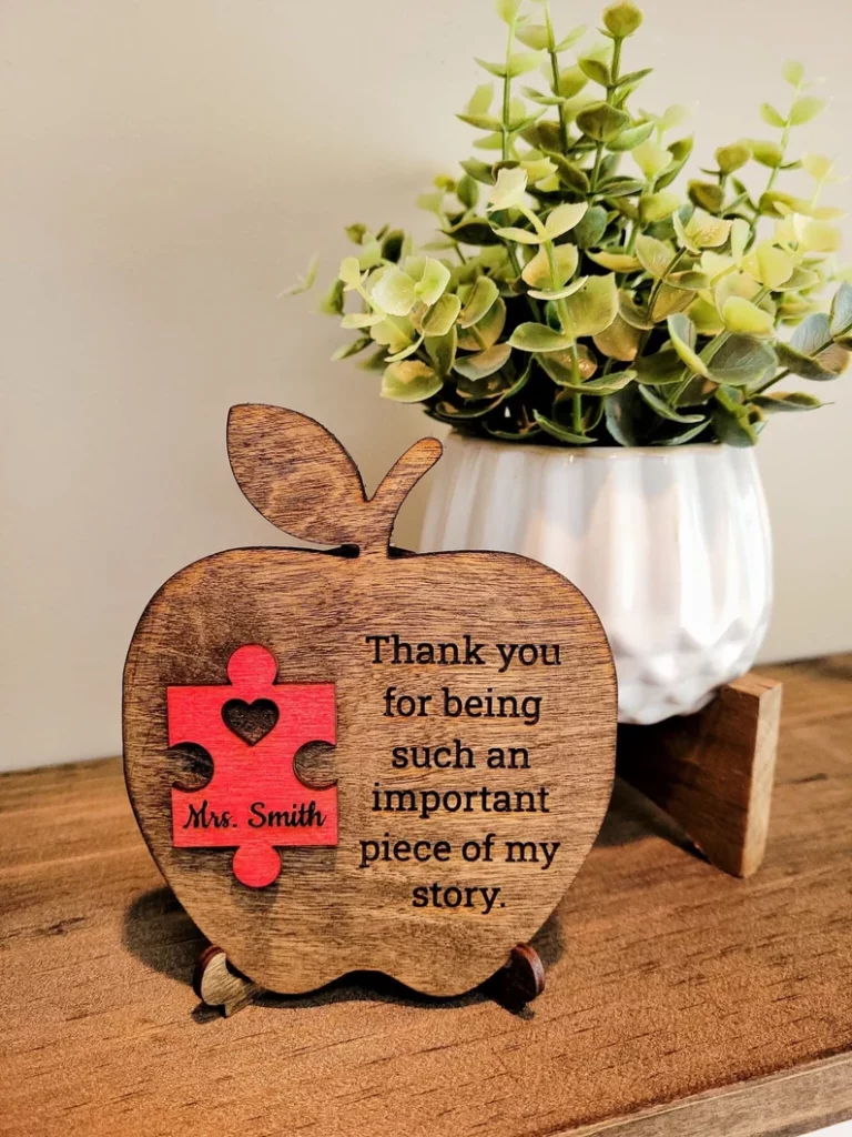 A wooden apple is engraved with a thank you note to a teacher. It has a red puzzle piece on it that has a teacher's name on it.