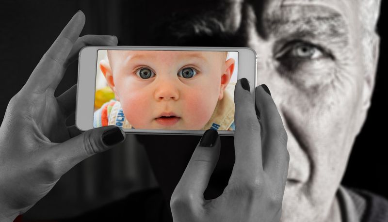 Hands holding up a phone with a picture of a baby's face in front an old man's face
