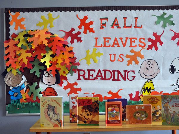 Fall bulletin boards uses puns like this one reading "fall leaves us reading." There are fall leaves and a tree as well as Linus, Peppermint Patty, Charlie Brown, and Snoopy. A table is in front of the board with Autumn themed books on it.