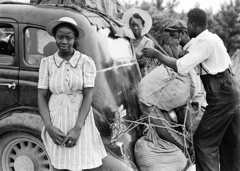 Vintage photo of a Black family strapping luggage to a car, with a young girl posing in front