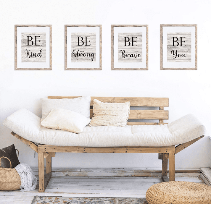 Four picture frames with the sayings "Be Kind," "Be Strong," "Be Brave," and "Be You" on a wall above a wooden bench with white cushions and pillows.