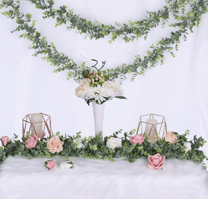 Floral garland hanging in a bunting style on a wall with a table featuring more garland, candles, and a bouquet of flowers in a vase below it.