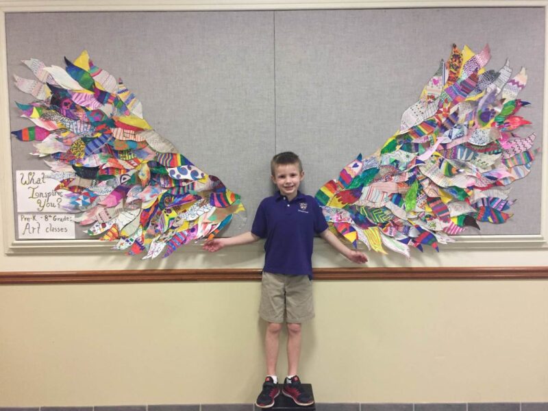 A little boy stands in front of a bulletin board with wings sticking out on either side of him. 