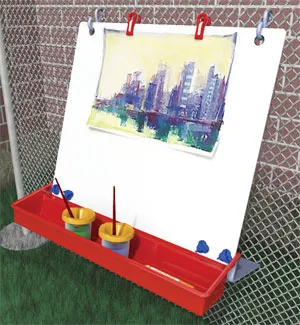 An easel is seen hanging from a wire fence outside in this example of an art easel for kids. A red tray holds cups of paint.