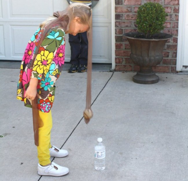 Student wearing a pantyhose leg hanging off their head with a ball at the end of it, trying to knock over a water bottle.