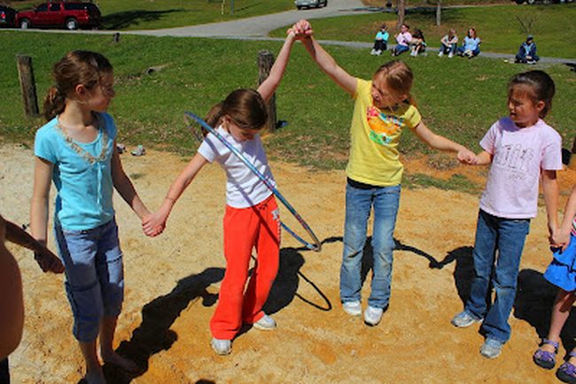 Students playing field day games are holding hands work to pass a hula hoop along the line without breaking the chain, stepping in and out of the hoop at it goes.