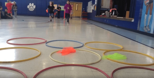 Rows of hula hoops laid out on a gym floor for field day games. Students are racing to drop their colored scarves into the hoops before the other team, trying to get three in a row.