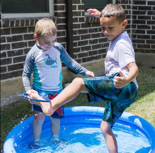 Two boys standing in a kiddie pool of water, trying to fish out items using their toes.