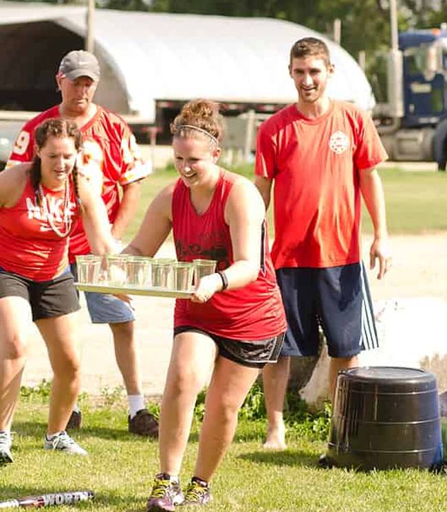 Woman tries to run carrying a tray of water glasses as her teammates watch.