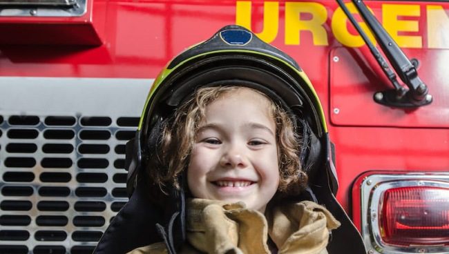 Preschooler wearing a fire helmet and standing in front of a fire truck (Field Trip Ideas)