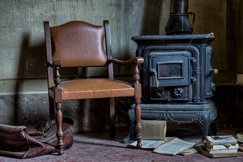 A leather chair next to an old woodstove with a valise and old books