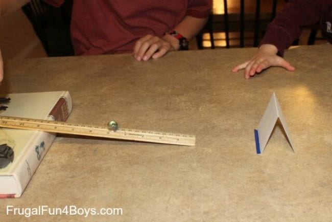 A ruler propped on one side on a flat book, with a marble rolling down it toward a folded index card (First Grade Science Experiments)