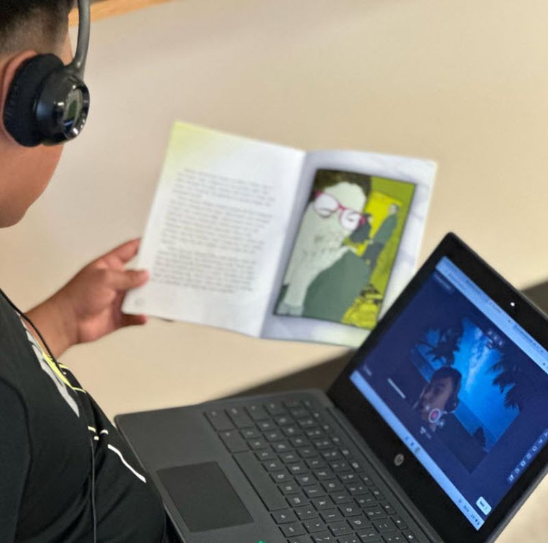 Student holding a book and wearing headphones, recording a video of themselves reading aloud using Flip on a laptop