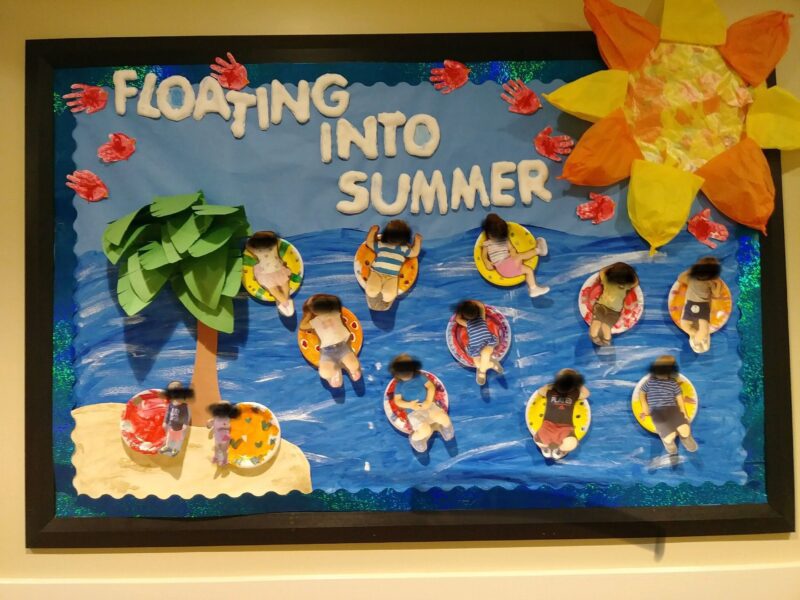 A bulletin board says Floating Into Summer. It has people floating on rafts in an ocean. A palm tree, sun, and sand are also included (summer bulletin boards ideas)