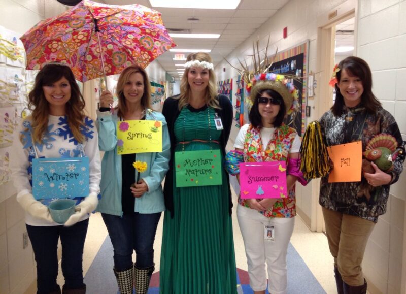 A woman in the middle is wearing a green dress with a sign that says mother nature. A woman dressed as winter and another dressed as spring are standing to her left. A woman to the right is dressed as summer and another woman to her right is dressed as fall.