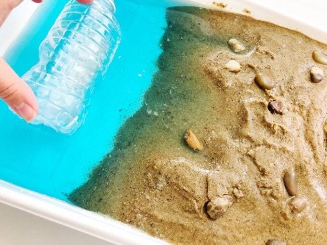 Plastic bin filled with sand, shells, and water to simulate a beach, with a hand holding a plastic bottle in the water (Fourth Grade Science)