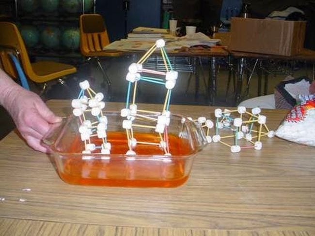 Fourth grade science teacher's hand shaking a pan of Jello topped with a house model made of toothpicks and marshmallows