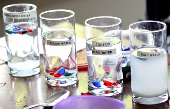 Series of glasses filled with liquid labeled baking soda water, sugar water, control plain water, and salt water, with red and blue objects floating in each