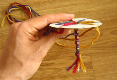 A hand is seen holding a small circle of cardboard with slits cut into it. Different colored yarn are shown thread through the slits and being braided beneath.